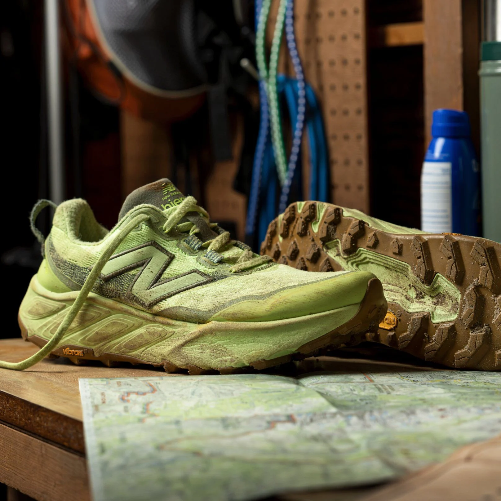 Pair of light green sneakers shown dusty and worn on a wooden workbench, with one shoe upright and the other flipped to display the sole, set beside a folded trail map. 